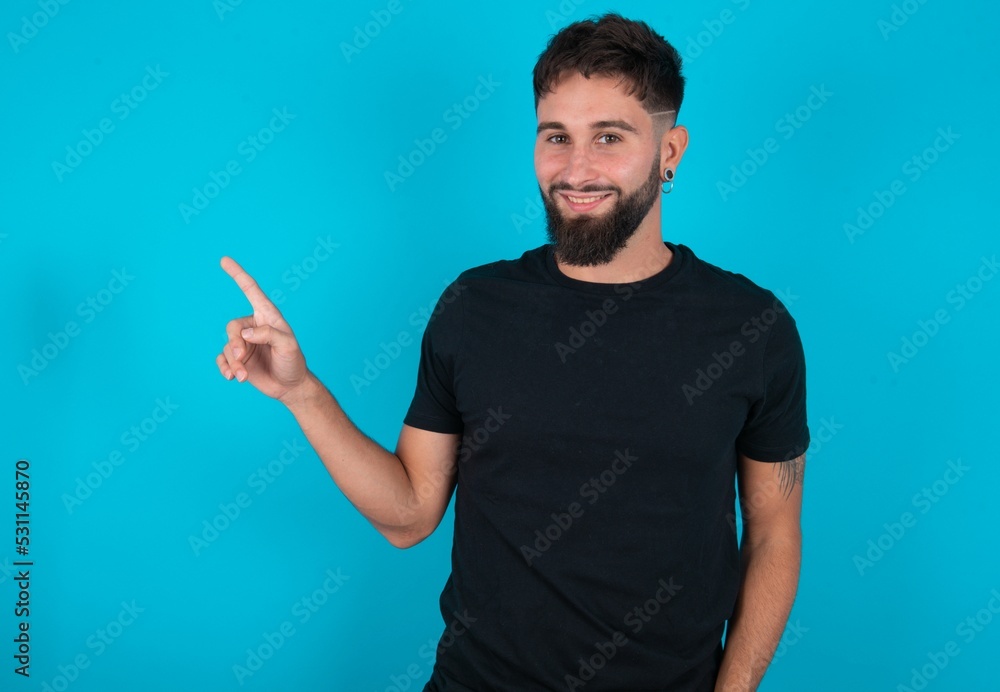 young hispanic bearded man wearing black T-shirt standing against blue background looking at camera indicating finger empty space sales young hispanic bearded man wearing black T-shirt standing against blue background looking at camera indicating finger empty space sales
