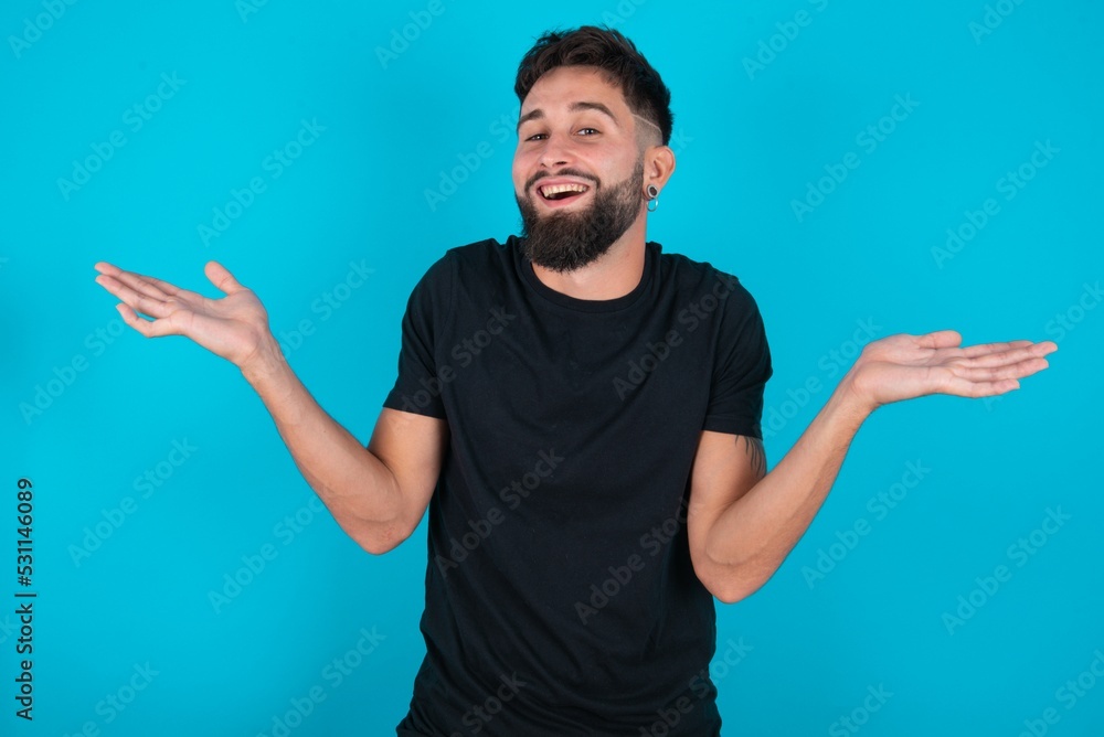 young hispanic bearded man wearing black T-shirt standing against blue background celebrating crazy and amazed for success with arms raised and open eyes screaming excited. Winner concept young hispanic bearded man wearing black T-shirt standing against blue background celebrating crazy and amazed for success with arms raised and open eyes screaming excited. Winner concept
