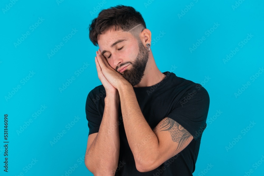 young hispanic bearded man wearing black T-shirt standing against blue background sleeping tired dreaming and posing with hands together while smiling with closed eyes. young hispanic bearded man wearing black T-shirt standing against blue background sleeping tired dreaming and posing with hands together while smiling with closed eyes.