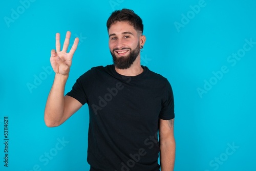 young hispanic bearded man wearing black T-shirt standing against blue background showing and pointing up with fingers number three while smiling confident and happy.