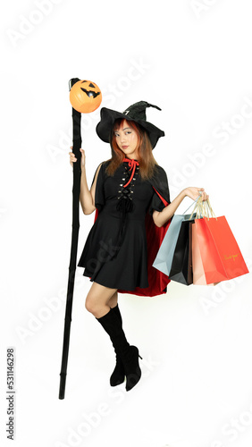 Beautiful girl with black and orange hair in black dress and witch hat is holding staff with shopping bags on white background for Halloween day.