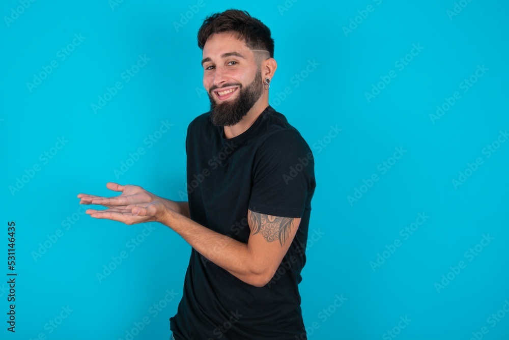 young hispanic bearded man wearing black T-shirt standing against blue background pointing aside with hands open palms showing copy space, presenting advertisement smiling excited happy young hispanic bearded man wearing black T-shirt standing against blue background pointing aside with hands open palms showing copy space, presenting advertisement smiling excited happy