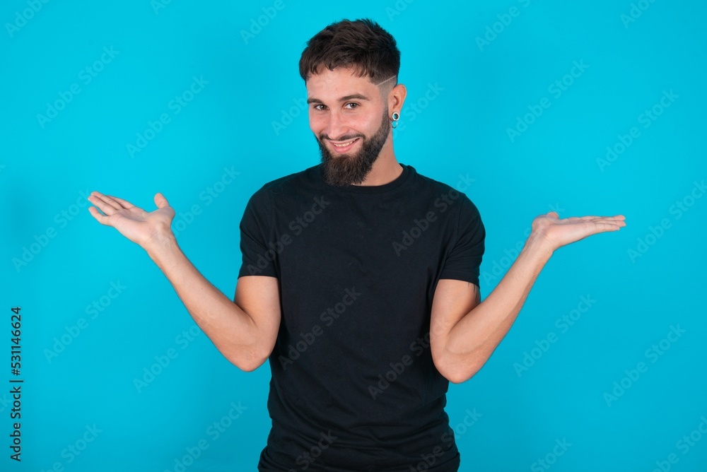 Cheerful cheery optimistic young bearded hispanic man wearing black T-shirt over blue background holding two palms copy space Cheerful cheery optimistic young bearded hispanic man wearing black T-shirt over blue background holding two palms copy space