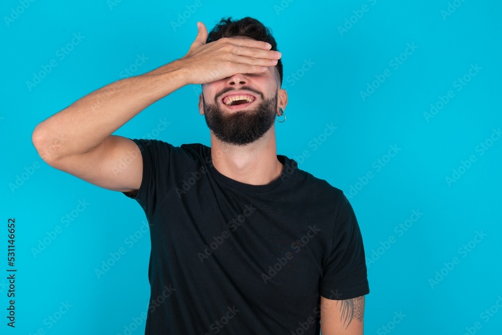 young bearded hispanic man wearing black T-shirt over blue background smiling and laughing with hand on face covering eyes for surprise. Blind concept. young bearded hispanic man wearing black T-shirt over blue background smiling and laughing with hand on face covering eyes for surprise. Blind concept.