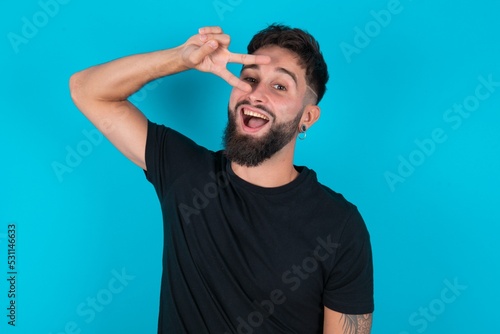 young bearded hispanic man wearing black T-shirt over blue background Doing peace symbol with fingers over face, smiling cheerful showing victory