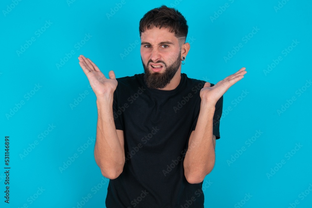 Indignant young bearded hispanic man wearing black T-shirt over blue background gestures in bewilderment, frowns face with dissatisfaction. Indignant young bearded hispanic man wearing black T-shirt over blue background gestures in bewilderment, frowns face with dissatisfaction.