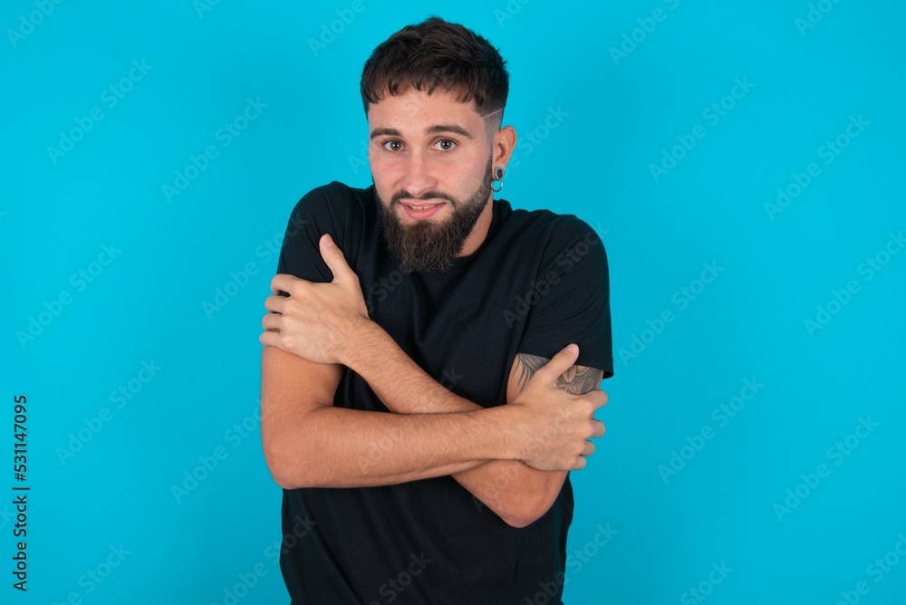 Desperate young bearded hispanic man wearing black T-shirt over blue background trembles and feels cold, hugs oneself to warm up or feels scared notices something terrifying. Desperate young bearded hispanic man wearing black T-shirt over blue background trembles and feels cold, hugs oneself to warm up or feels scared notices something terrifying.