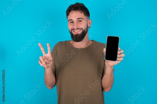 young handsome latin man wearing casual clothes standing over blue background holding modern device showing v-sign
