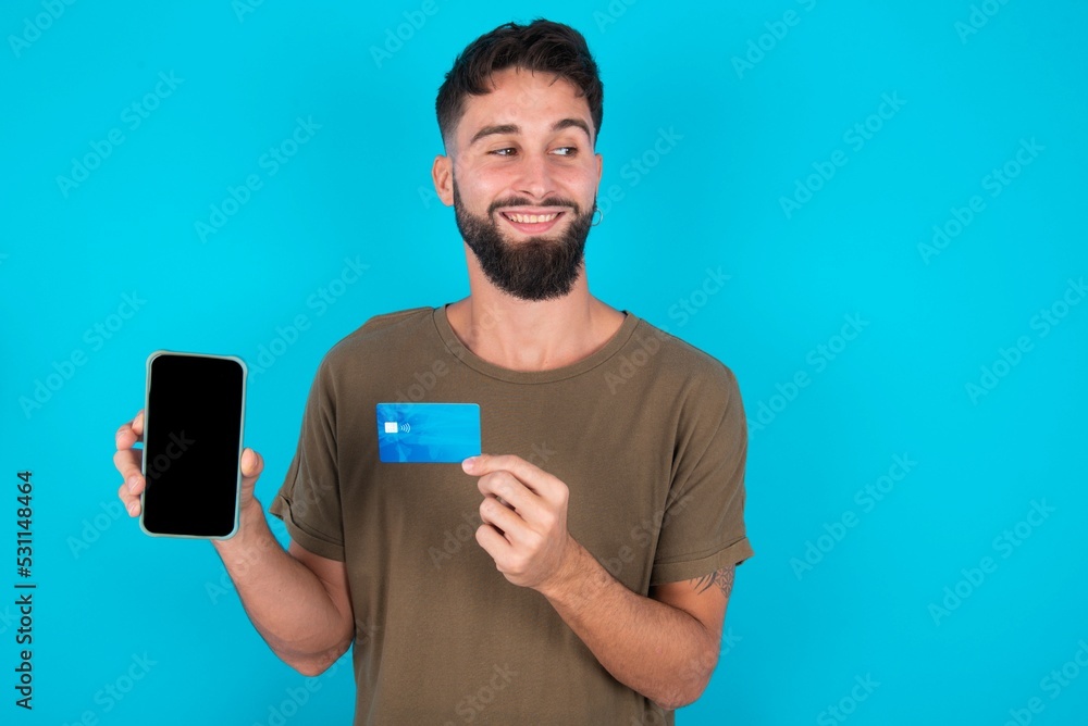 young handsome latin man wearing casual clothes standing over blue background holding bank card modern device looking empty space young handsome latin man wearing casual clothes standing over blue background holding bank card modern device looking empty space