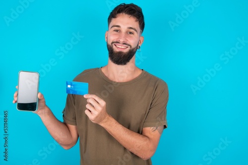 Photo of adorable young handsome latin man wearing casual clothes standing over blue background holding credit card and Smartphone. Reserved for online purchases