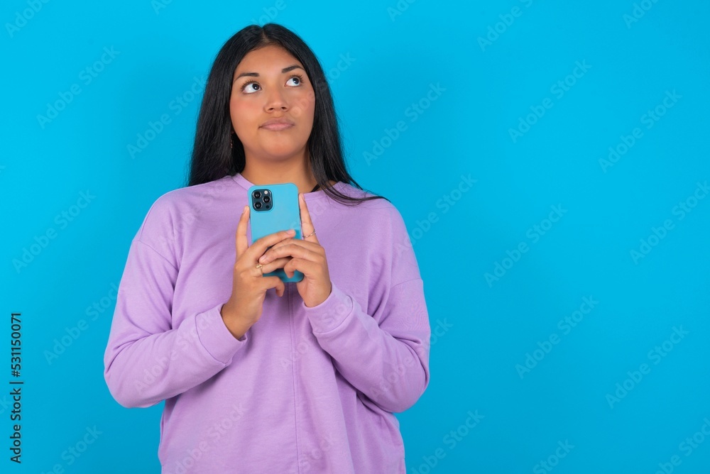 Portrait of young beautiful latin woman wearing casual clothes standing against blue background with dreamy look, thinking while holding smartphone. Tries to write up a message. Portrait of young beautiful latin woman wearing casual clothes standing against blue background with dreamy look, thinking while holding smartphone. Tries to write up a message.