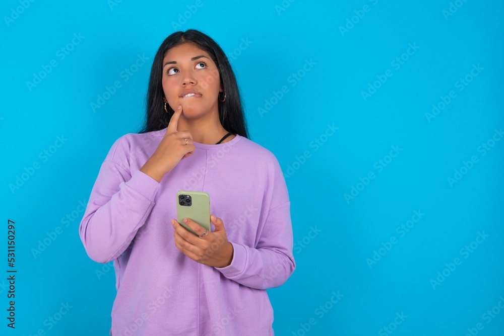 Image of a thinking dreaming young beautiful latin woman wearing casual clothes standing against blue background using mobile phone and holding hand on face. Taking decisions and social media concept. Image of a thinking dreaming young beautiful latin woman wearing casual clothes standing against blue background using mobile phone and holding hand on face. Taking decisions and social media concept.