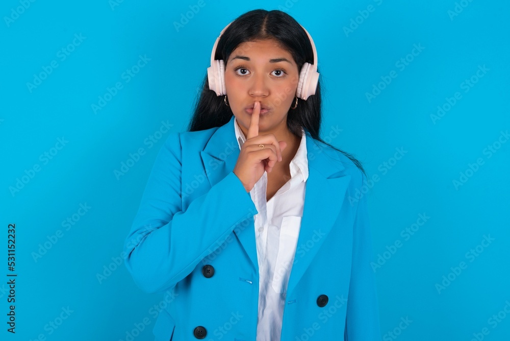 Young latin businesswoman wearing blue blazer over blue background making hush gesture with finger on her lips wearing wireless headphones. Be quiet. Young latin businesswoman wearing blue blazer over blue background making hush gesture with finger on her lips wearing wireless headphones. Be quiet.