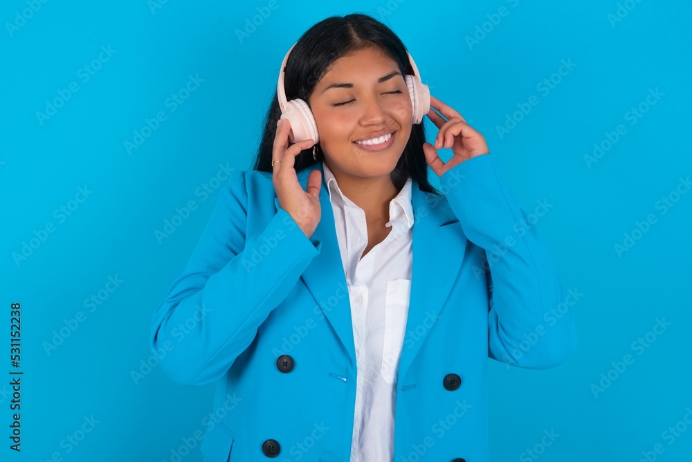 Young latin businesswoman wearing blue blazer over blue background with headphones on head, listens to music, enjoying favourite song with closed eyes, holding hands on headset. Young latin businesswoman wearing blue blazer over blue background with headphones on head, listens to music, enjoying favourite song with closed eyes, holding hands on headset.