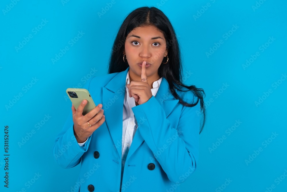 Young latin businesswoman wearing blue blazer over blue background holding modern gadget ask not tell secrets Young latin businesswoman wearing blue blazer over blue background holding modern gadget ask not tell secrets