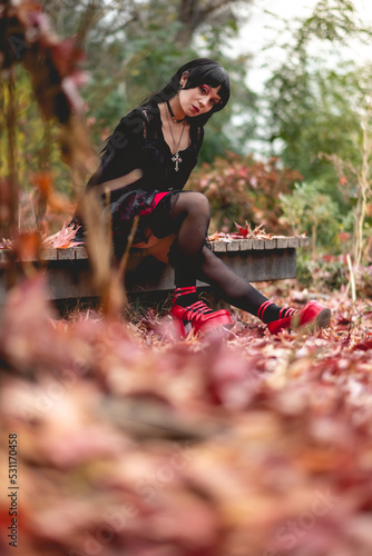 Fototapeta Naklejka Na Ścianę i Meble -  Young and skinny hispanic goth girl with black dress and red platforms shoes seated in a wood platform in the autumn forest with red, orange and yellow dry leaves