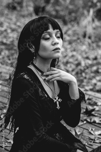 Young and skinny hispanic goth girl with black dress, cross, sexy face and makeup seated in a wood platform in the autumn forest with dry leaves (in black and white) © Samuel Ponce