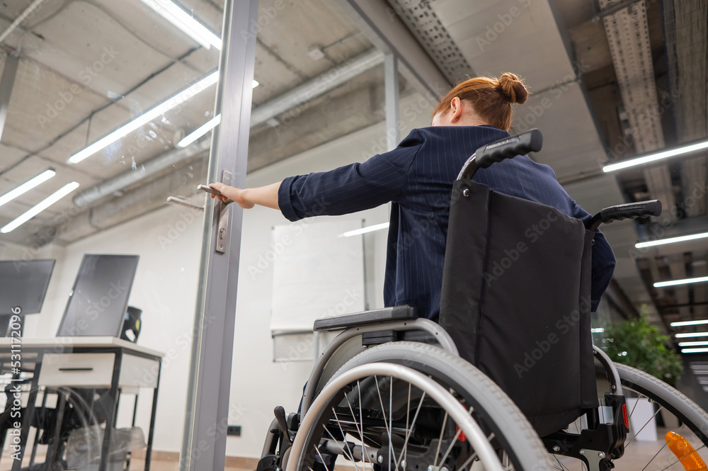 Red-haired caucasian woman in a wheelchair trying to open the door in the office. Red-haired caucasian woman in a wheelchair trying to open the door in the office.