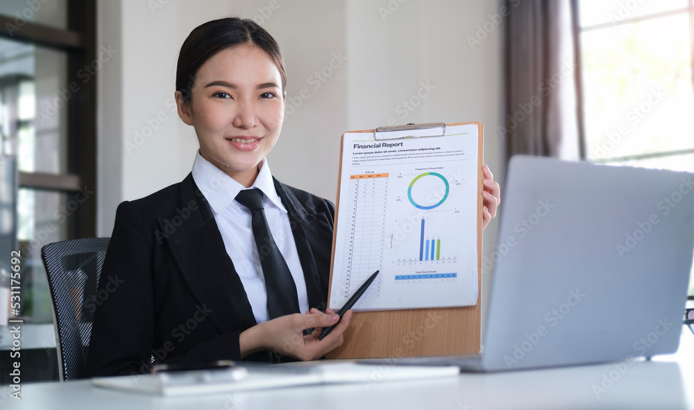 Confident asian woman in business suit sitting front of laptop computer and showing financial document to camera. Confident asian woman in business suit sitting front of laptop computer and showing financial document to camera.