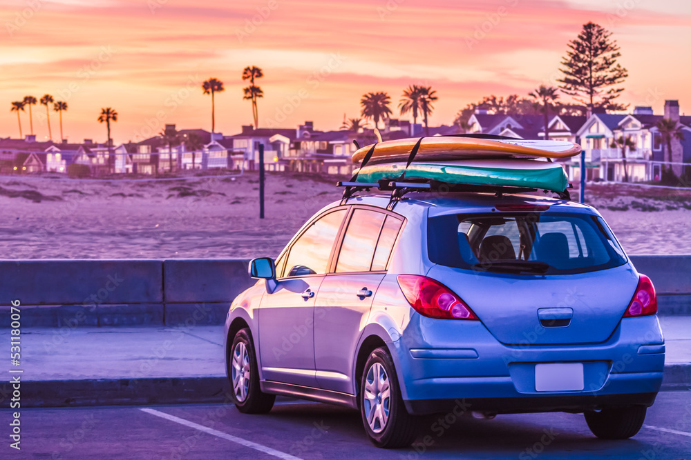 A blue car with the surfing board and longboard on the roof parked near the beach, lightened by the setting sun