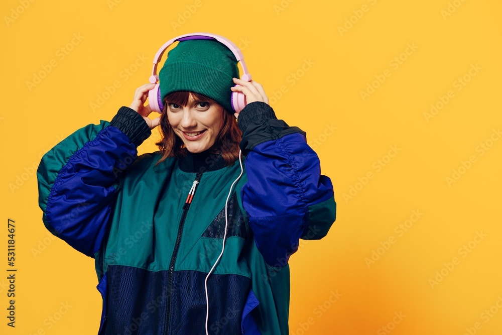 a happy, joyful woman in a green jacket and hat enjoys music standing in pink headphones on a yellow background holding them with her hands and looking at the camera a happy, joyful woman in a green jacket and hat enjoys music standing in pink headphones on a yellow background holding them with her hands and looking at the camera