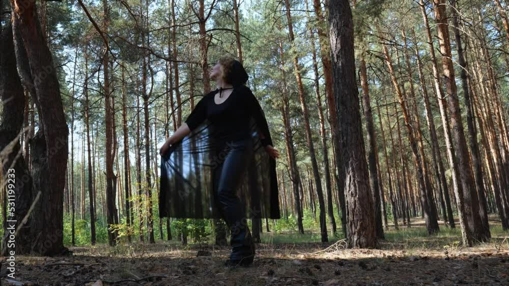 An adult woman in a black cloak with a hood dances in a pine forest. Halloween woman moving. woman magic in nature An adult woman in a black cloak with a hood dances in a pine forest. Halloween woman moving. woman magic in nature