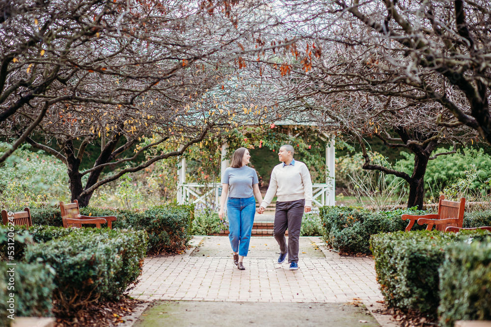 smiling lgbt couple holding hands with dead trees, shrubs and wooden benches on the side smiling lgbt couple holding hands with dead trees, shrubs and wooden benches on the side