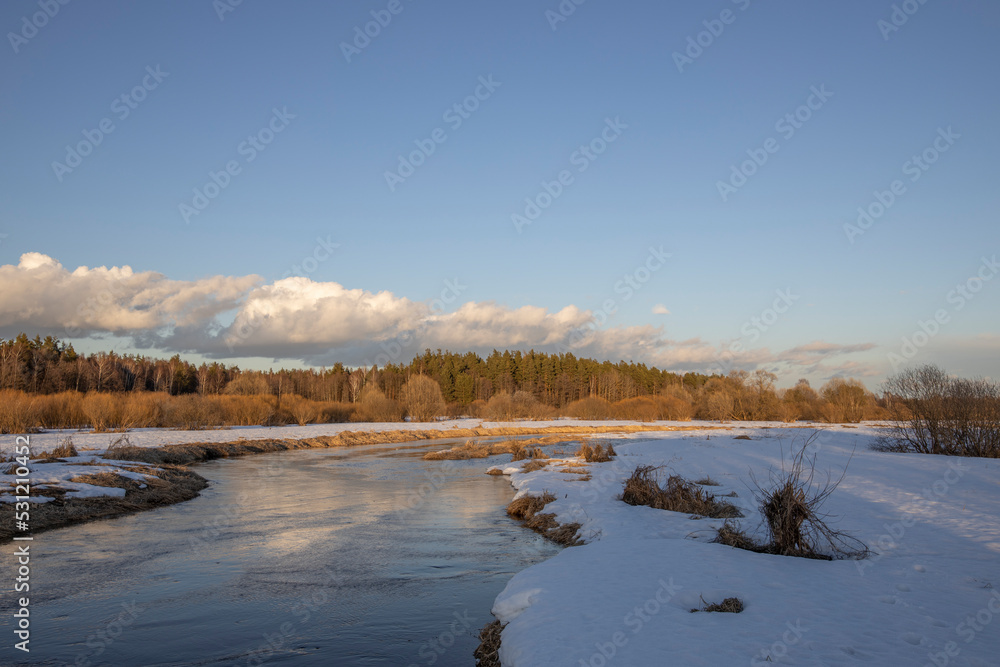 March sunny evening by the river. Blue sky over the horizon. A picturesque landscape, early spring, a river with snow-covered banks, dry grass and bushes. The first thaws, the snow is melting March sunny evening by the river. Blue sky over the horizon. A picturesque landscape, early spring, a river with snow-covered banks, dry grass and bushes. The first thaws, the snow is melting