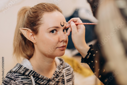 Make-up artist makes month moon on forehead face of young attractive Caucasian blonde woman. Woman model with extended elf ears looking at camera. Halloween face art painting, lifestyle portrait
