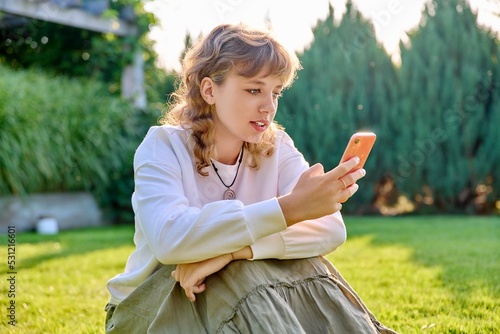 Cute teenage girl sitting with smartphone on grass in backyard