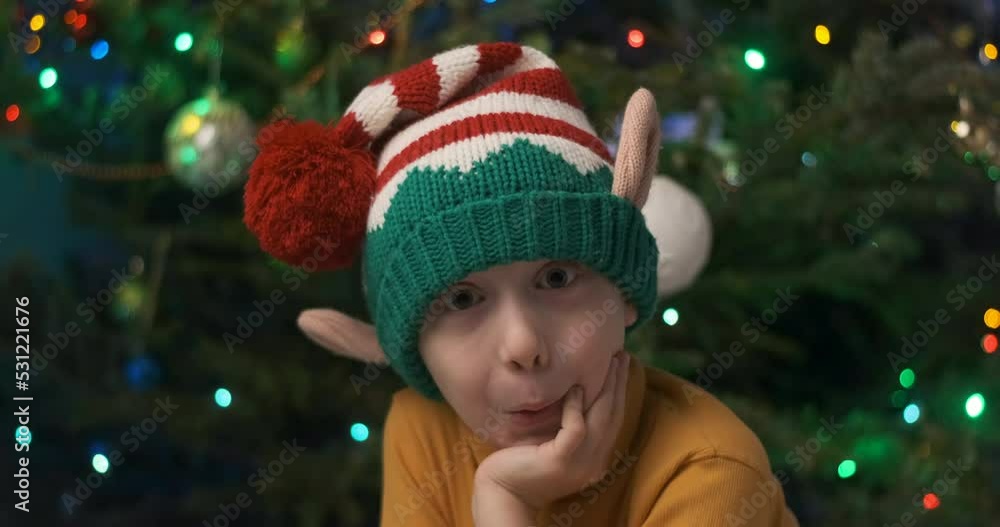 Boy in knitted gnome hat under Christmas tree and garland background. Video portrait of brooding boy on Christmas Eve. Boy in knitted gnome hat under Christmas tree and garland background. Video portrait of brooding boy on Christmas Eve.