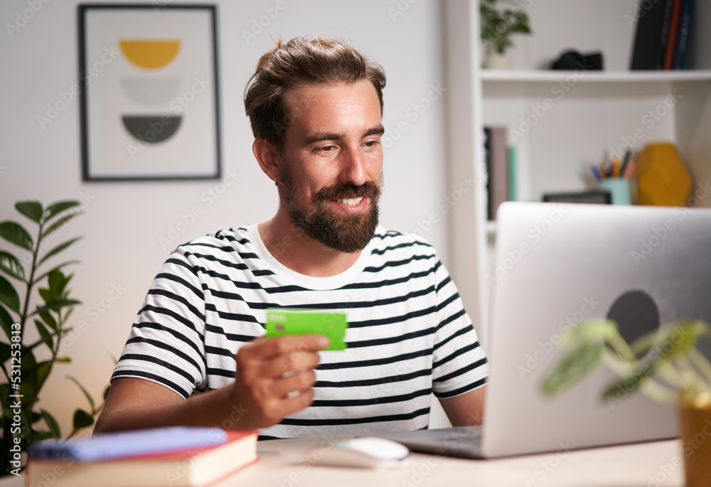 Cheerful young man doing e-shopping using laptop at home. Happy caucasian blond male making online payments with credit card for service or goods. Self employed freelancer collecting fee. Shopaholic. Cheerful young man doing e-shopping using laptop at home. Happy caucasian blond male making online payments with credit card for service or goods. Self employed freelancer collecting fee. Shopaholic.