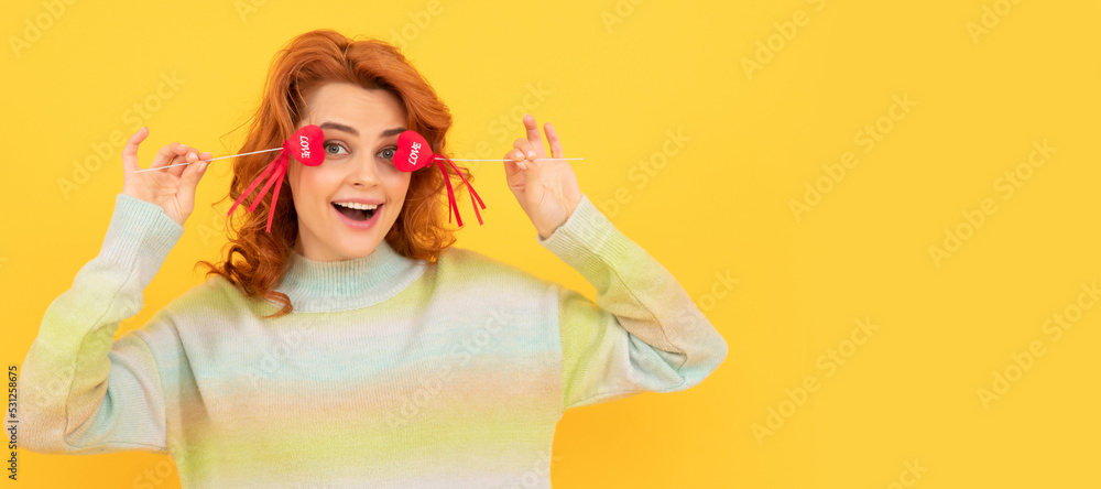 glad funny redhead girl with red heart sticks on yellow background. february 14. Woman isolated face portrait, banner with copy space.