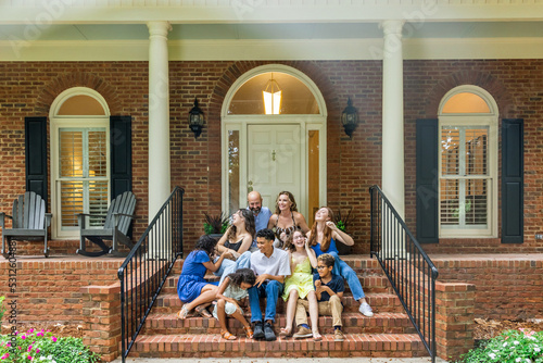 A large and blended and mixed happy family sitting on the front steps of their brick home in the suburbs © Ursula Page