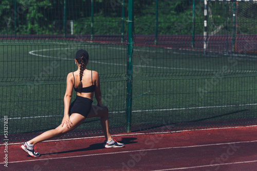 young woman doing deep lunge stretching on sports ground outdoors in summer.