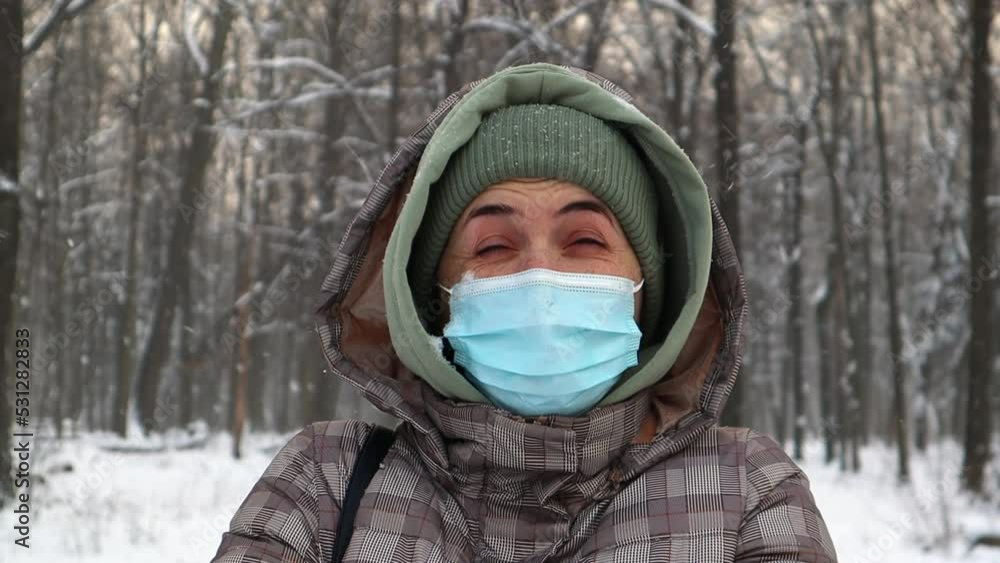 Happy woman in a protective medical mask rejoices in the snowfall outdoors in a city park in winter against the backdrop of trees. Positive female woman rejoices in winter nature and white snow. Happy woman in a protective medical mask rejoices in the snowfall outdoors in a city park in winter against the backdrop of trees. Positive female woman rejoices in winter nature and white snow.