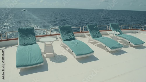 The raw of daybeds on the upper deck of a moving luxurious diving safari yacht with small coffee tables in between and an ocean horizon in the background; several deck chairs on the top of a ship