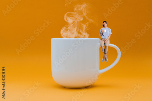 Huge Cup of hot coffee and woman sitting on mug handle and drinking coffee isolated on yellow background. Creative idea for coffee time