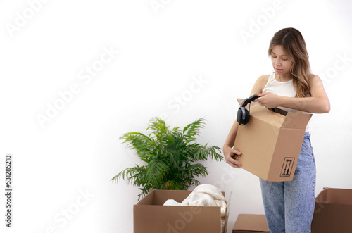 A young woman holds a box in her hands and takes out headphones. Nearby are boxes with things and a flower in a pot. Moving to a new house. High quality photo