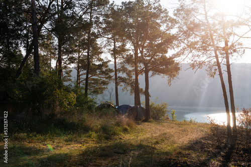 Distance view. Morning landscape. Pine forest, sun rays, glow, river lake, mountains. Summer camping warm colors. Tourism and vacation.