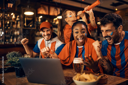 Fotografie, Obraz Group of excited sports fans celebrating their favorite team's victory while watching match on laptop in pub