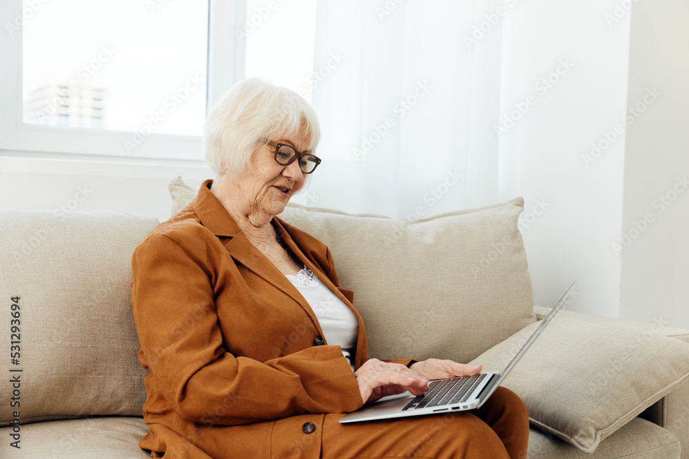 a radiant, successful elderly woman in a stylish brown pantsuit sits on a spacious sofa with a laptop on her lap and smiles happily, covering her eyes with pleasure a radiant, successful elderly woman in a stylish brown pantsuit sits on a spacious sofa with a laptop on her lap and smiles happily, covering her eyes with pleasure