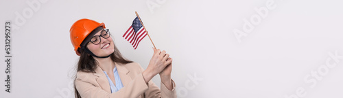 A girl in a construction helmet and an American flag stands happy on a white background.