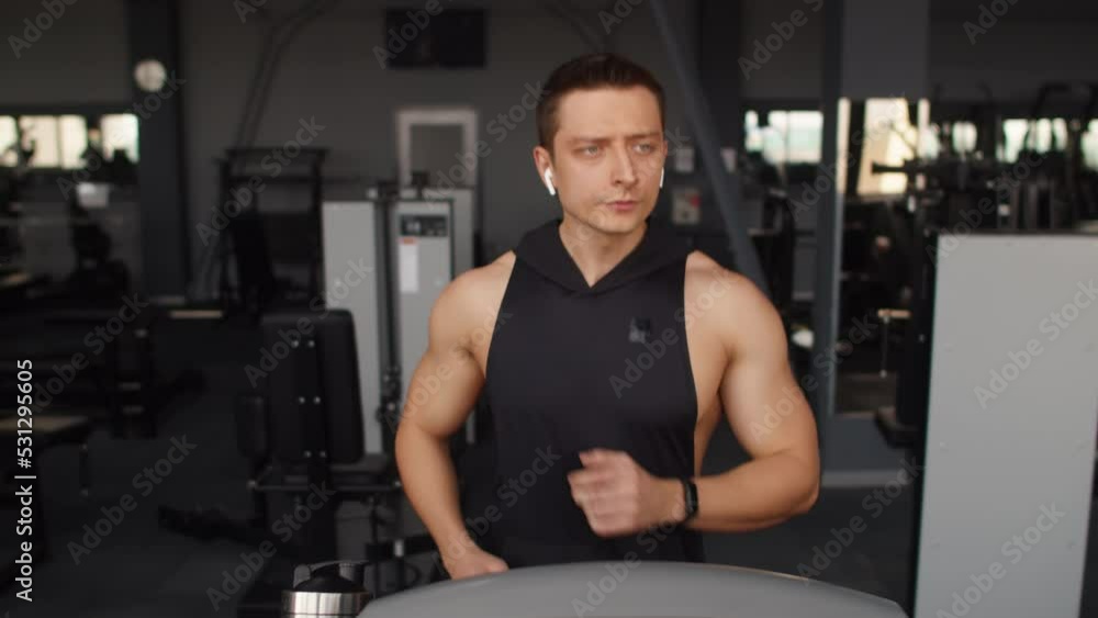 Handsome athletic man in black sportswear running on a treadmill at the gym, tapping his white wireless earbuds headphones, listening to music or answering phone call Handsome athletic man in black sportswear running on a treadmill at the gym, tapping his white wireless earbuds headphones, listening to music or answering phone call