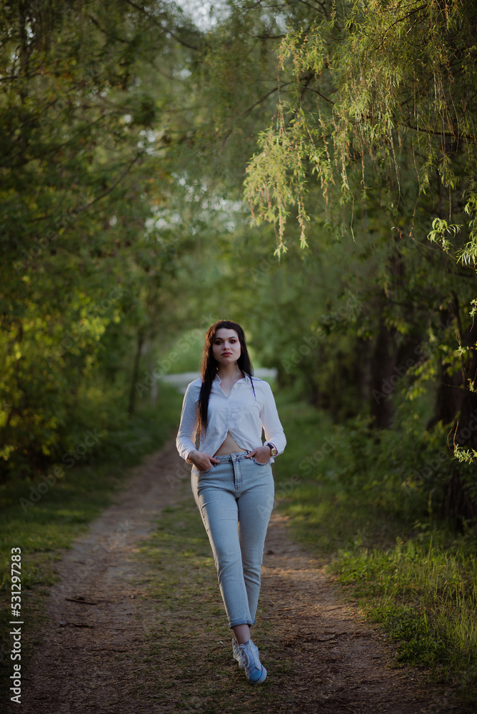 Sexy sensual brunette walks in a summer forest at sunset in a white blouse and jeans Sexy sensual brunette walks in a summer forest at sunset in a white blouse and jeans