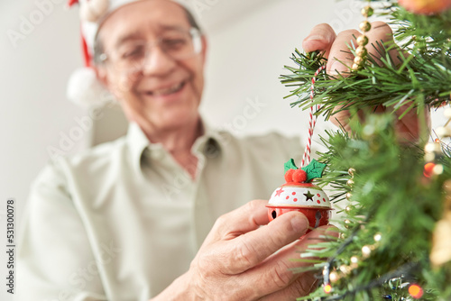 Happy senior hispanic man smiling while decorating a Christmas tree at home wearing a red Santa Claus beanie. Selective focus on the foreground, the hands hanging a Christmas ball from a branch.