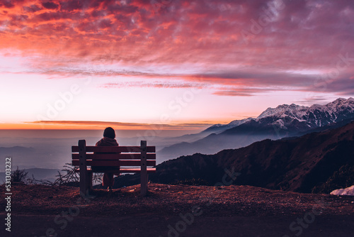 Silhouette of a Girl sitting on bench at Sunset - Girl looking at Snow Capped Mountains with Orange Scattered Clouds in the Sky - Winter Season - anime style concept