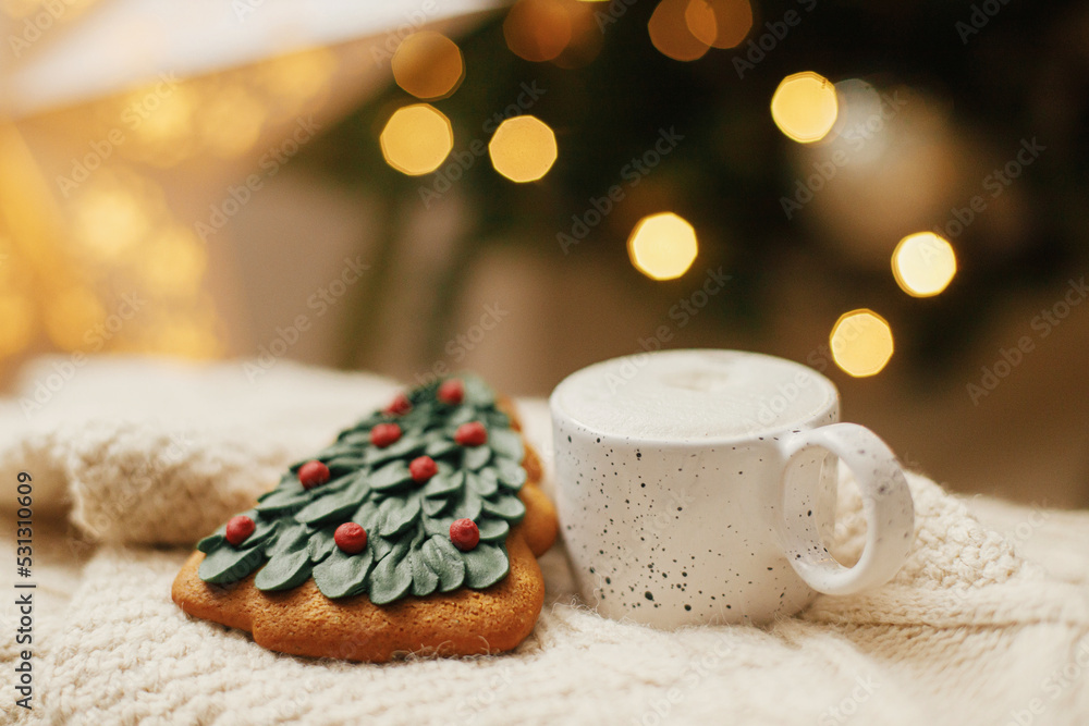 Fototapeta premium Atmospheric winter hygge. Warm cup of coffee and christmas tree gingerbread cookie on knitted sweater on background of christmas tree with lights. Christmas banner. Cozy holidays
