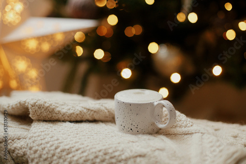 Warm cup of coffee on background of christmas tree with lights. Cozy home. Atmospheric winter hygge. Stylish mug on knitted sweater against golden illumination bokeh. Christmas banner