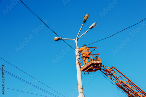 A municipal worker in protective equipment performs hazardous work to eliminate an interruption in the power grid. A worker repairs a street lamp from an aerial platform basket. photo